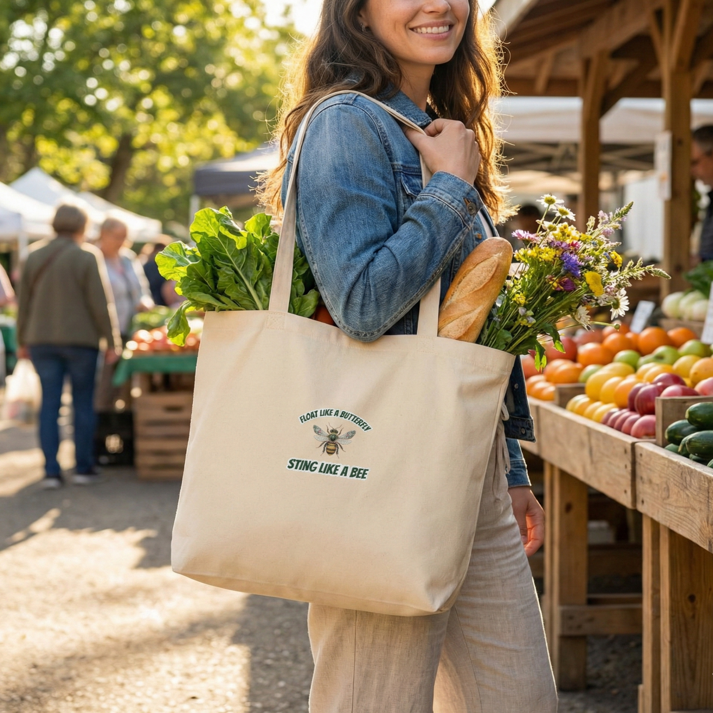 A lifestyle mockup of a woman at a sunlit outdoor farmers' market, carrying the oyster-colored "Float like a butterfly, sting like a bee" tote bag over her shoulder. The bag is filled with fresh greens, a baguette, and wildflowers, while wooden crates of colorful produce like apples and oranges line the stalls in the background.