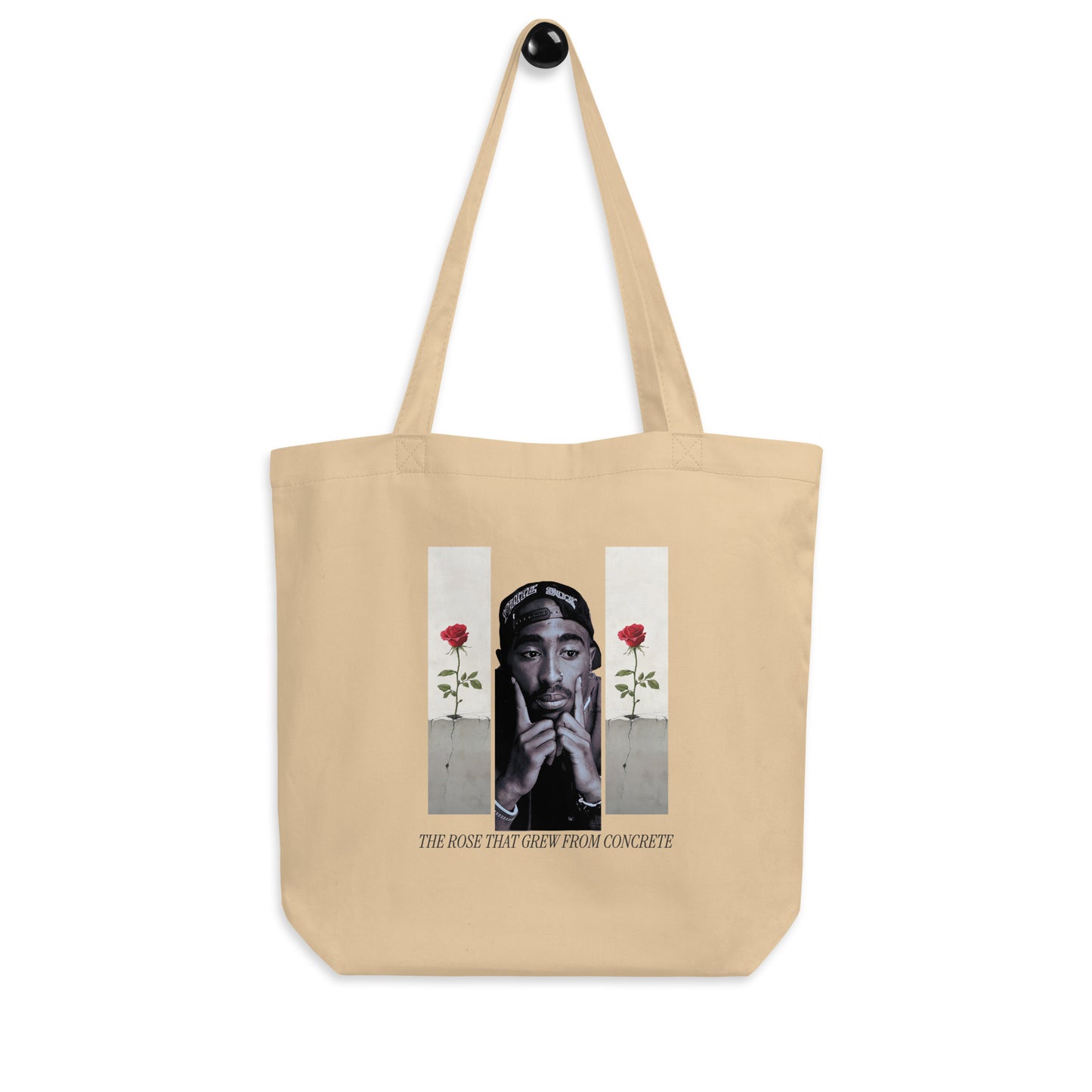 A beige canvas tote bag with a printed graphic of Tupac Shakur in the center, flanked by two vertical panels with red roses, and the text 'THE ROSE THAT GREW FROM CONCRETE' below. The bag is shown hanging from a black hook against a plain white background.