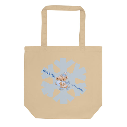 A beige canvas tote bag with a cartoon teddy bear wearing a snow hat and scarf, standing in front of a large light blue snowflake. The text 'catching vibes and snowballs' is written in blue around the bear.