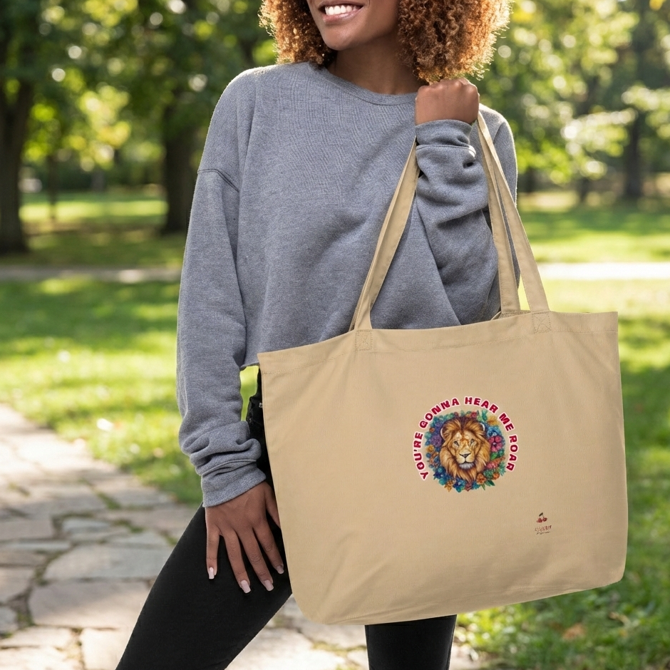 person with curly hair, wearing a grey sweatshirt and black pants, smiles while holding a light brown tote bag over their shoulder. The bag features a circular graphic with a lion's head and a colorful mane, surrounded by the red text "YOU'RE GONNA HEAR ME ROAR." They are standing on a stone path in a park with green grass and trees in the background.