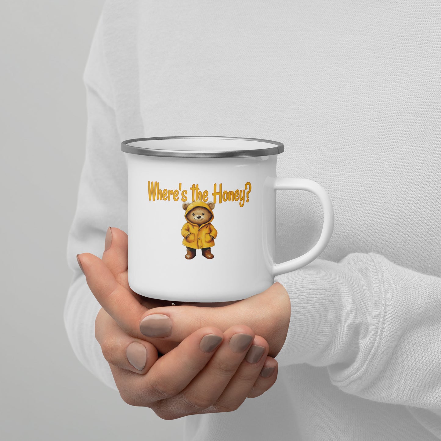 A person in a white sweatshirt holds a white enamel-style mug with a silver rim. The mug has a yellow text reading 'Where's the Honey?' and a small cartoon bear wearing a yellow raincoat.