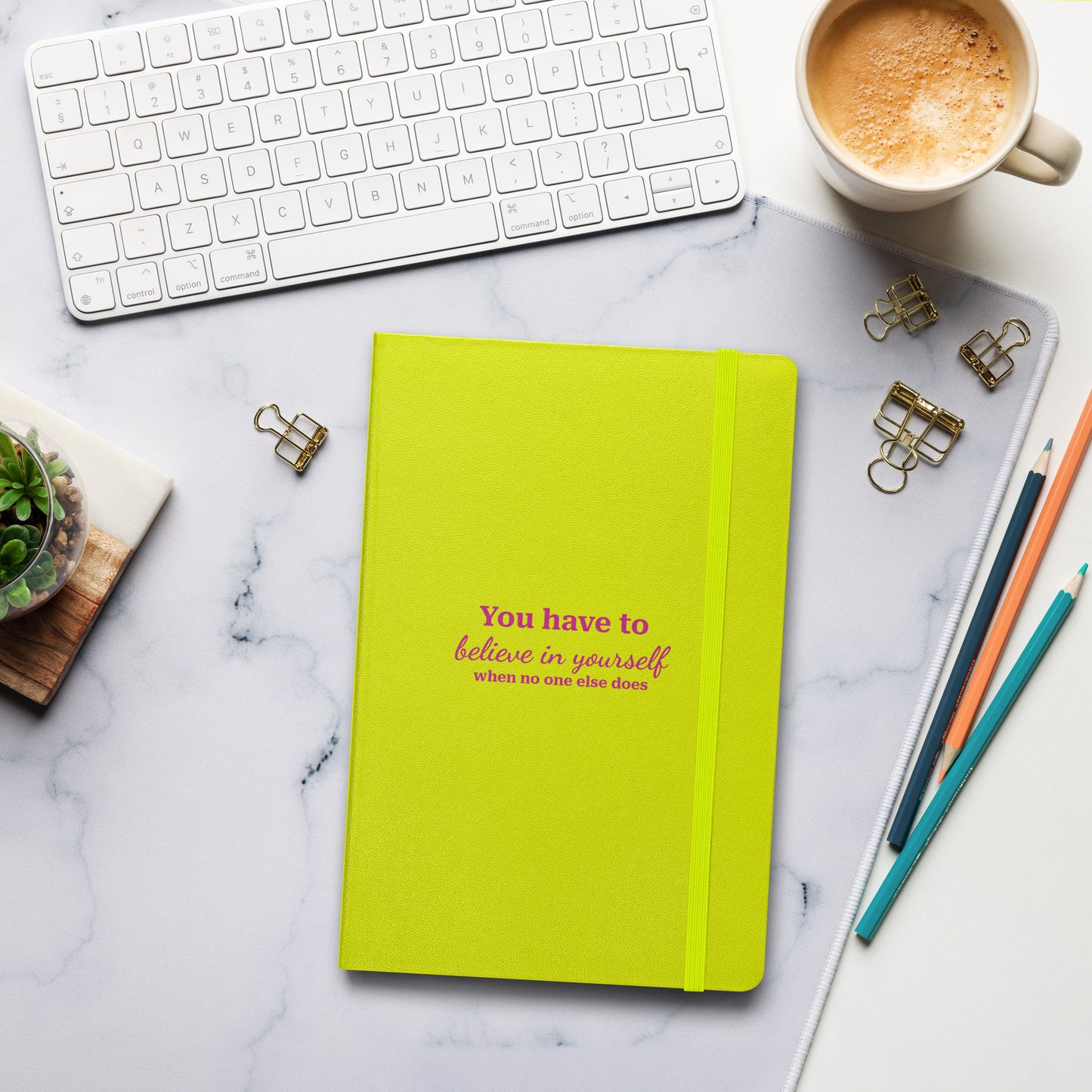 Lime green hardcover notebook with the text "You have to believe in yourself" on a marble desk with a keyboard and coffee.
