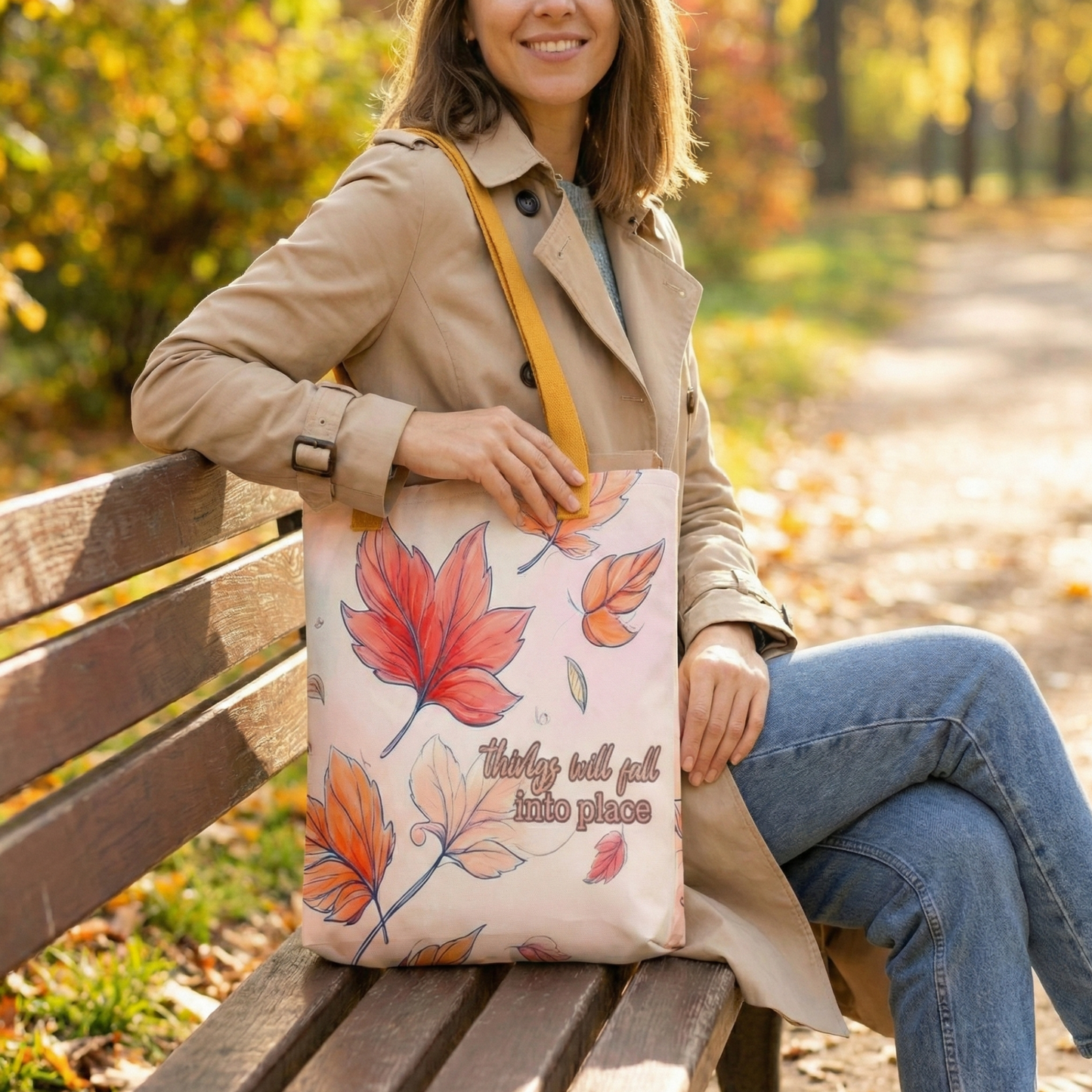 A woman sitting on a bench holding a tote bag with autumn leaf design and with the text "Thing will fall into place" in a park which has early autumn vibes.