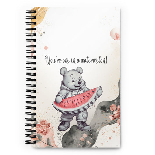 A spiral-bound notebook with a white cover featuring a cartoon bear holding a watermelon slice and the text 'You're one in a watermelon!'. The cover has a watercolor-style design with pink flowers and a black spiral binding.