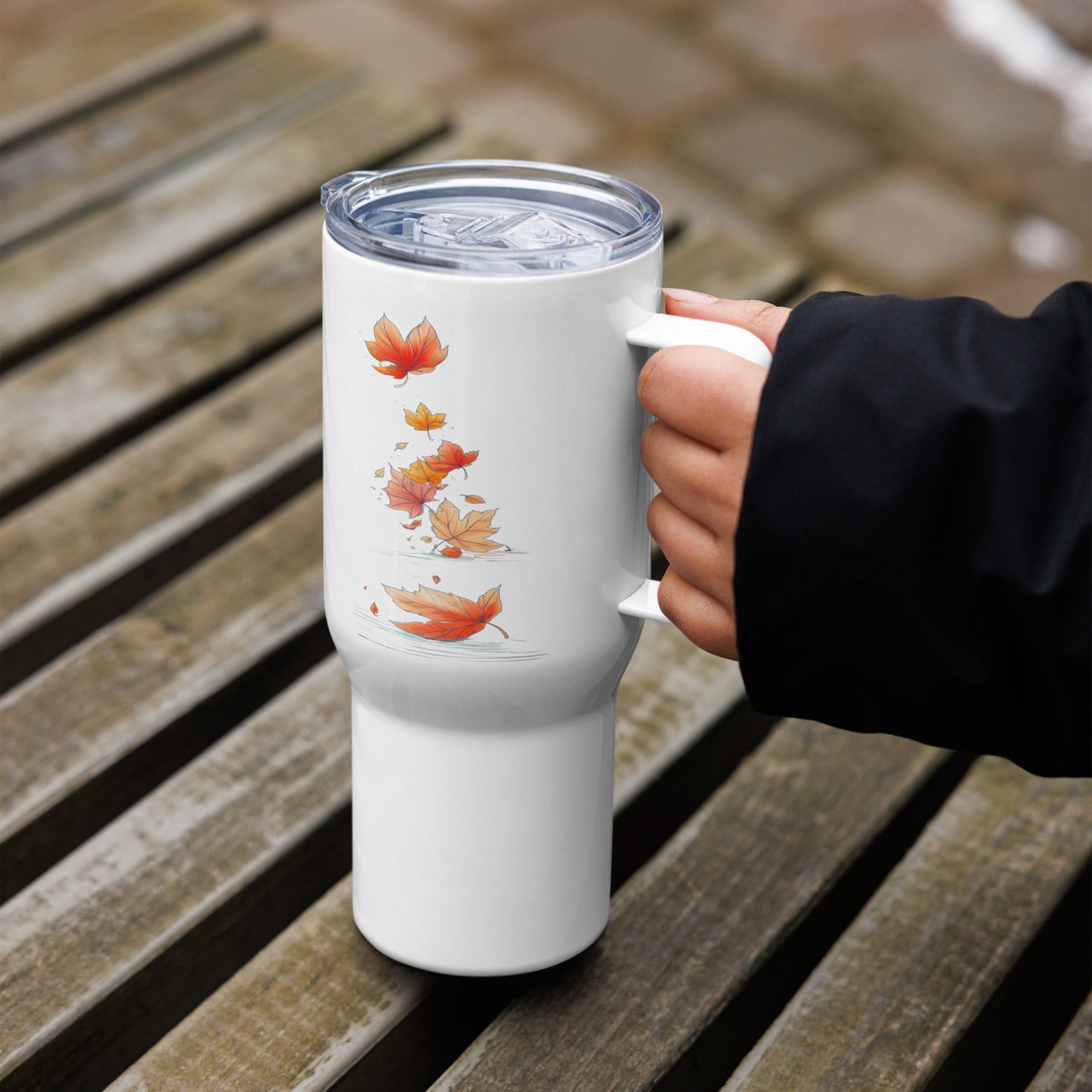 A white insulated travel tumbler with a clear lid and a printed design of orange and yellow autumn leaves, held by a hand on a wooden slatted surface.