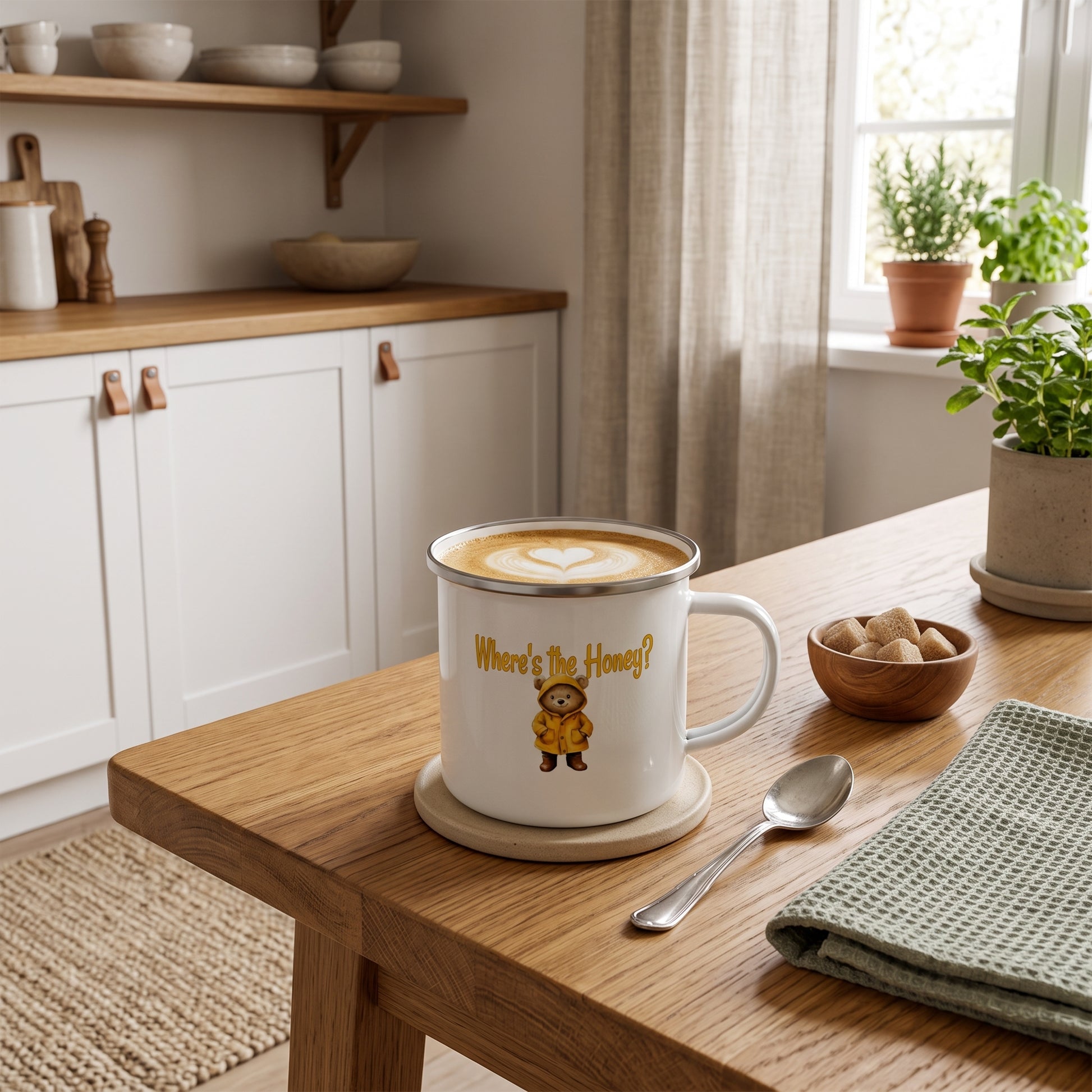 A white enamel mug with a silver rim sits on a wooden table in a bright kitchen. The mug features a cute cartoon bear wearing a yellow jacket and the text 'Where's The Honey? by Sweet & Savage'.