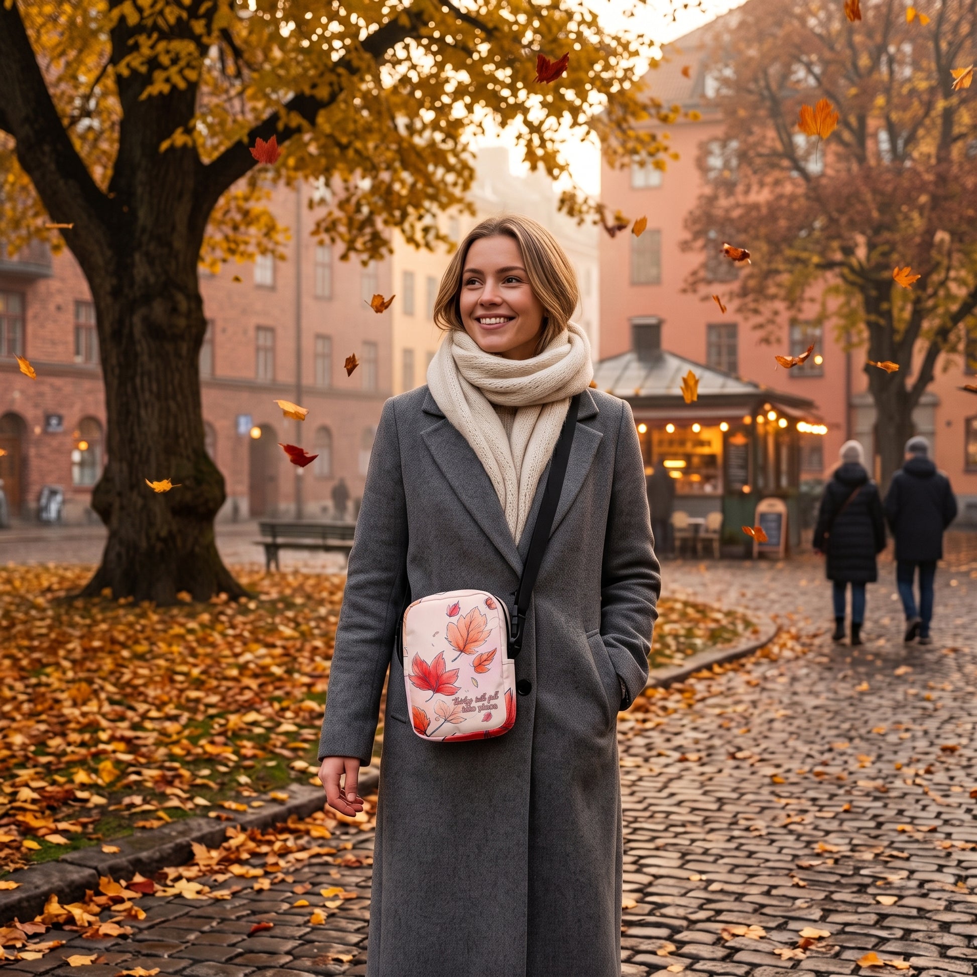 A woman in a gray coat and cream scarf stands on a cobblestone street in an autumn park, wearing a small crossbody bag with a red and white maple leaf pattern by Sweet & Savage. 