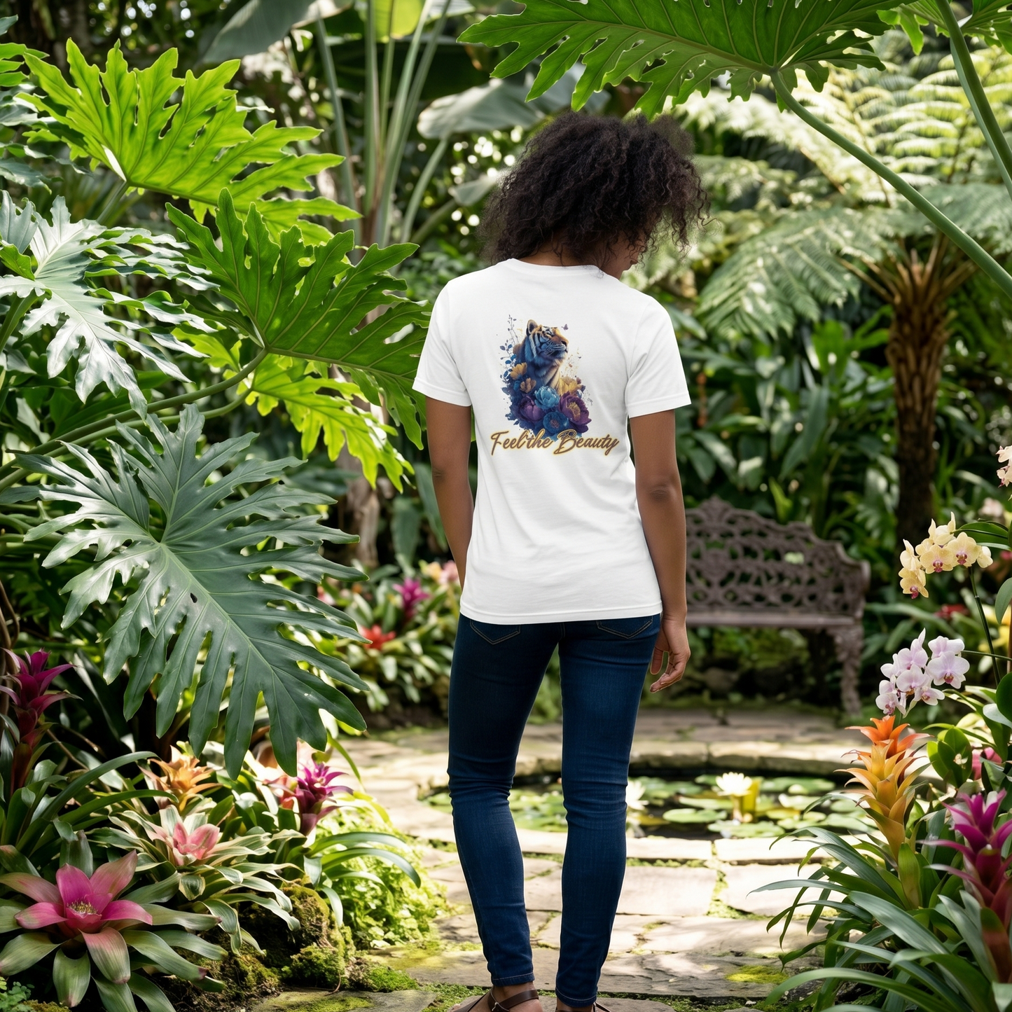 Back view of a white cotton t-shirt featuring a 'Feel the Beauty' tiger and floral illustration, worn by a woman walking through a lush botanical garden with tropical plants and a stone path.