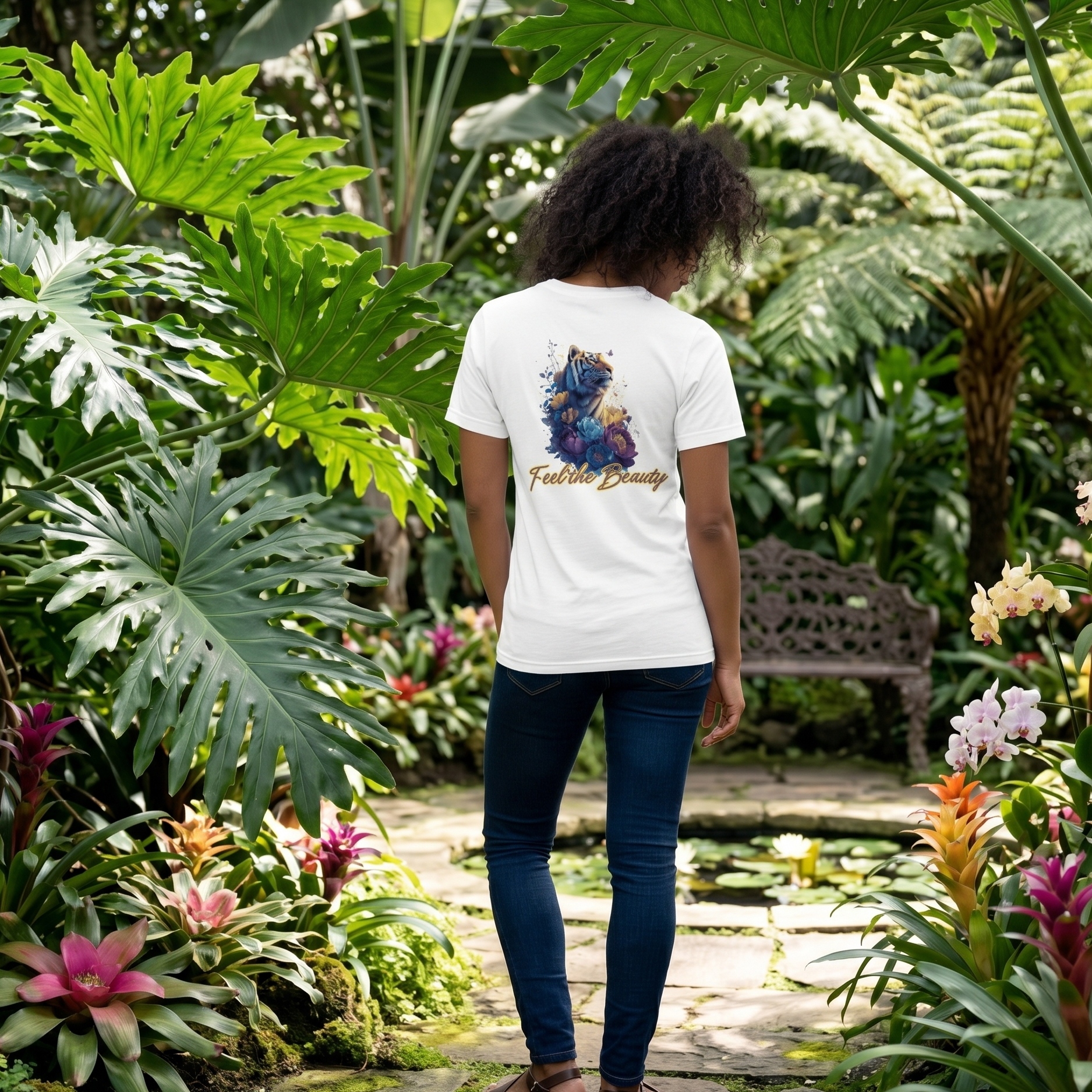 Back view of a white cotton t-shirt featuring a 'Feel the Beauty' tiger and floral illustration, worn by a woman walking through a lush botanical garden with tropical plants and a stone path.