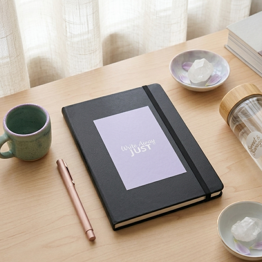 Mindful morning lifestyle mockup of 'Write Away JUST' black hardcover notebook with a lilac panel. Features the journal on a light wood desk next to a ceramic mug, a rose gold pen, white crystals, and a Sweet & Savage glass water bottle in soft natural light.