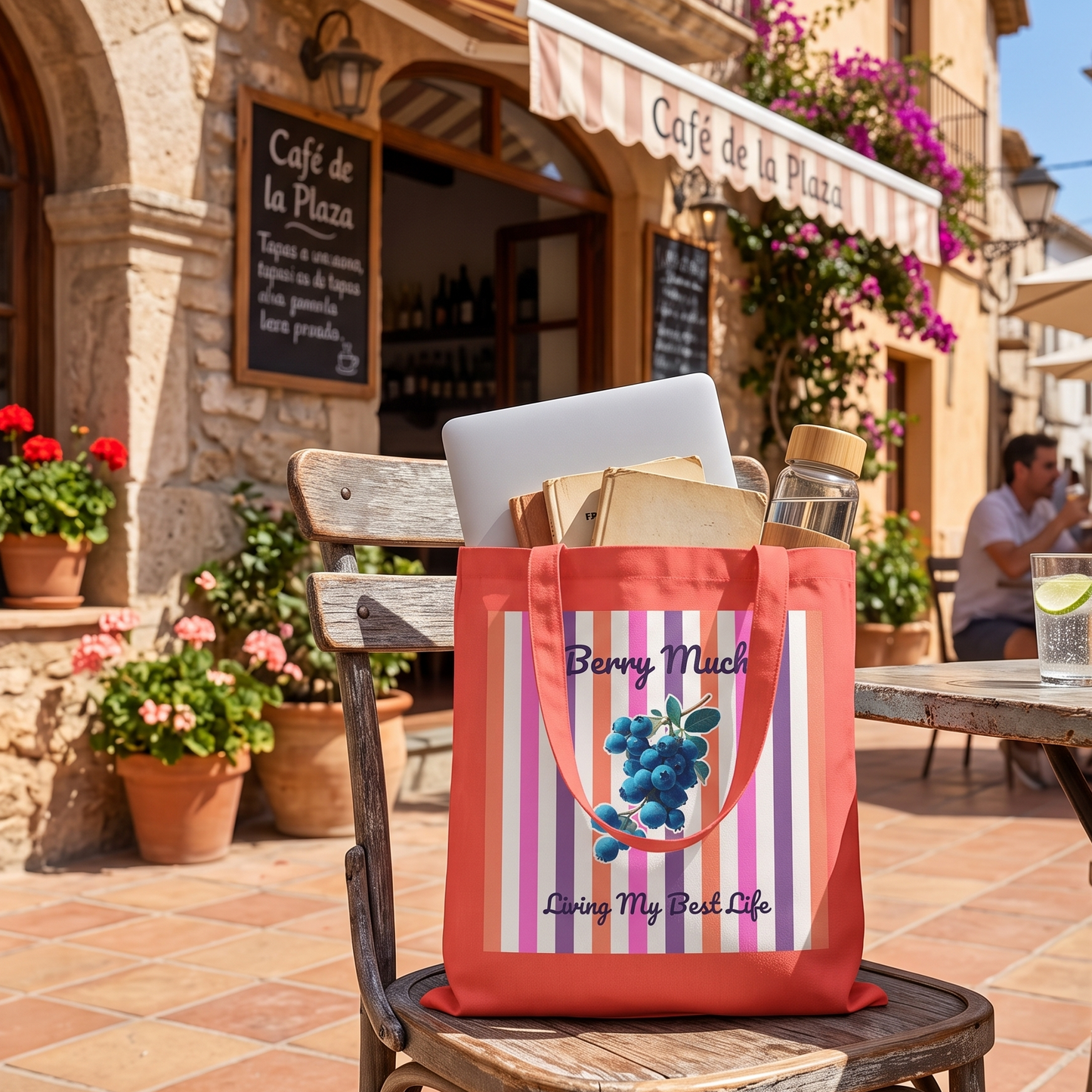 A coral canvas tote bag with purple and white vertical stripes, featuring a blueberry illustration and the text 'Berry Much' and 'Living My Best Life', sitting on a wooden chair in front of a cafe patio.