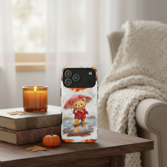 A MagSafe iPhone tough case with a "Magic of Autumn" watercolor bear design resting on a dark wood table. Beside the phone is a glowing amber candle, a stack of vintage books, and a miniature orange pumpkin. A soft, white knit blanket is draped in the background, creating a cozy hygge reading nook aesthetic.