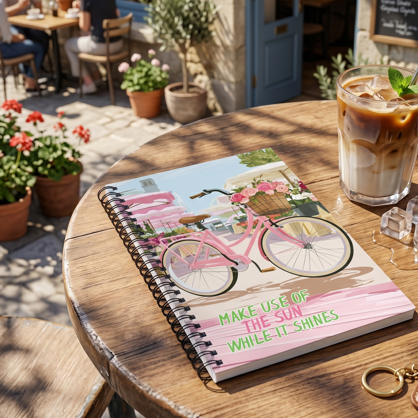 A spiral-bound notebook with a pink bicycle illustration and the text 'MAKE USE OF THE SUN WHILE IT SHINES' by Sweet & Savage lies on a round wooden table at an outdoor cafe, next to an iced coffee.
