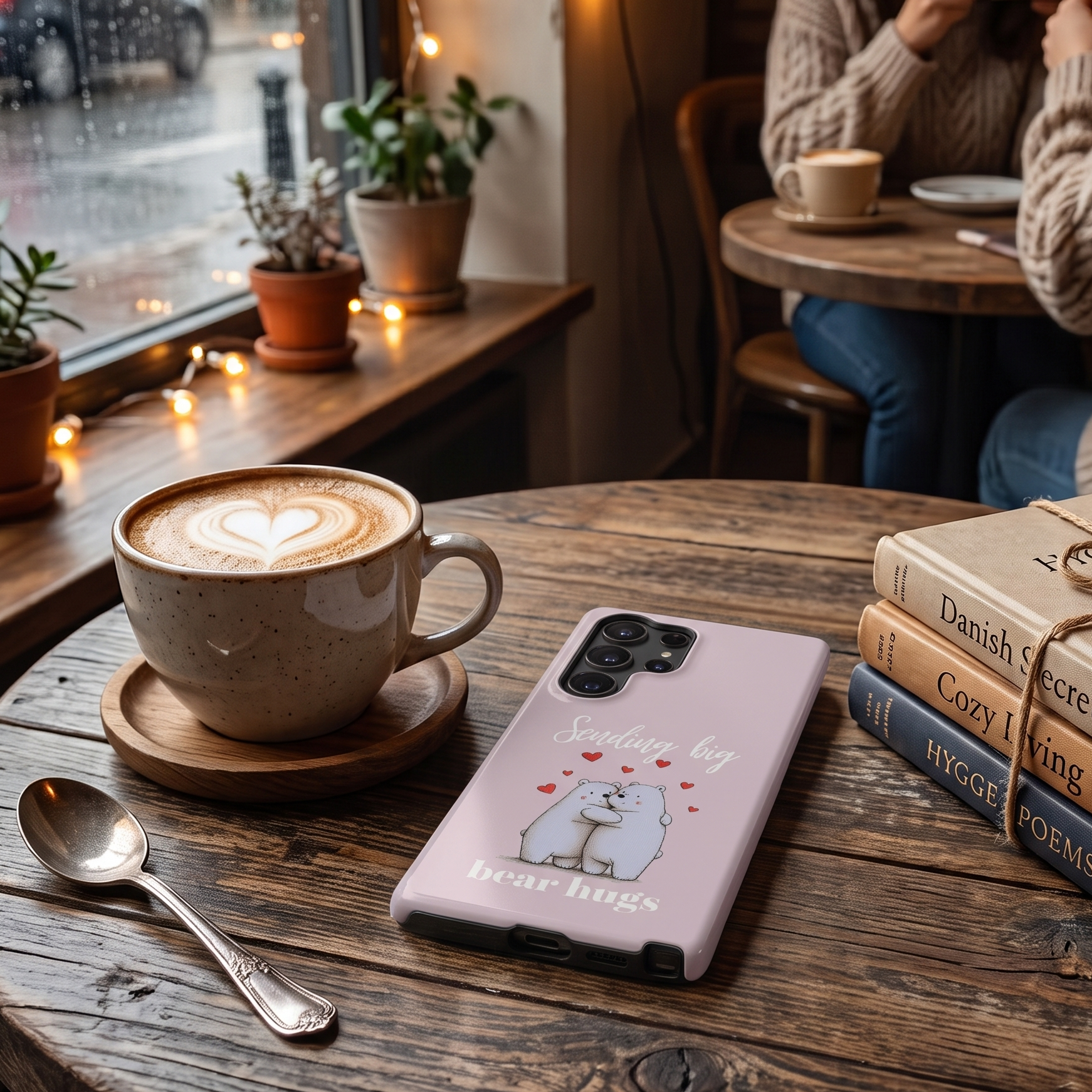 A light pink phone case featuring cute illustration of polar bears with the text 'Sending big bear hugs'' and small red hearts, resting on a rustic wooden table in a cozy cafe setting next to a cup of latte and books.