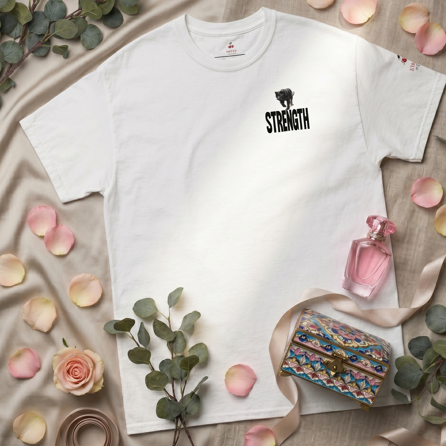 En hvid Sweet & Savage T-shirt i økologisk bomuld liggende fladt med et sort panter på brystet. Ved siden af ligger friske blomster og dekorative blade, der understreger brandets fokus på natur og bæredygtig mode.