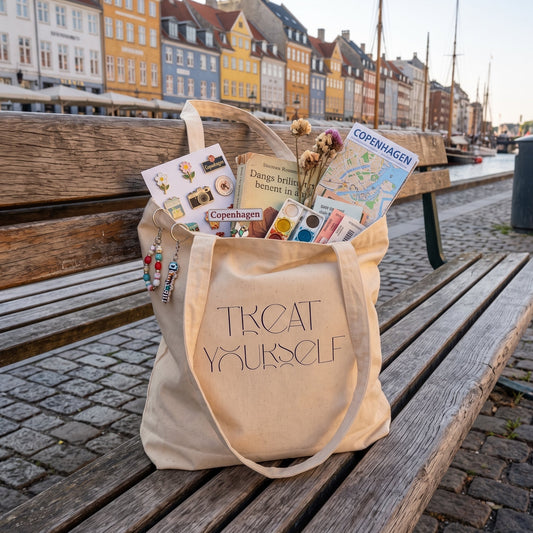 A beige canvas tote bag printed with 'TREAT YOURSELF' sits on a wooden bench in Nyhavn, Copenhagen, filled with travel-themed items including a map, postcards, and pins.
