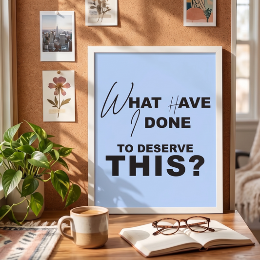 Framed poster with text 'What have I done to deserve this?' on a table with a cup, book, and glasses.