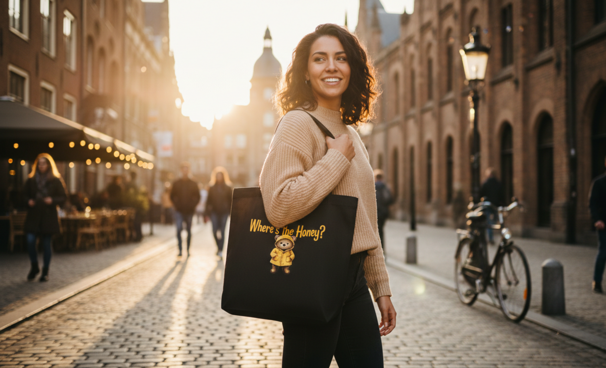 A smiling woman carrying a black eco-friendly tote bag featuring a whimsical bear in a yellow raincoat with the text 'Where's the Honey?', walking down a sunlit European-style cobblestone street.
