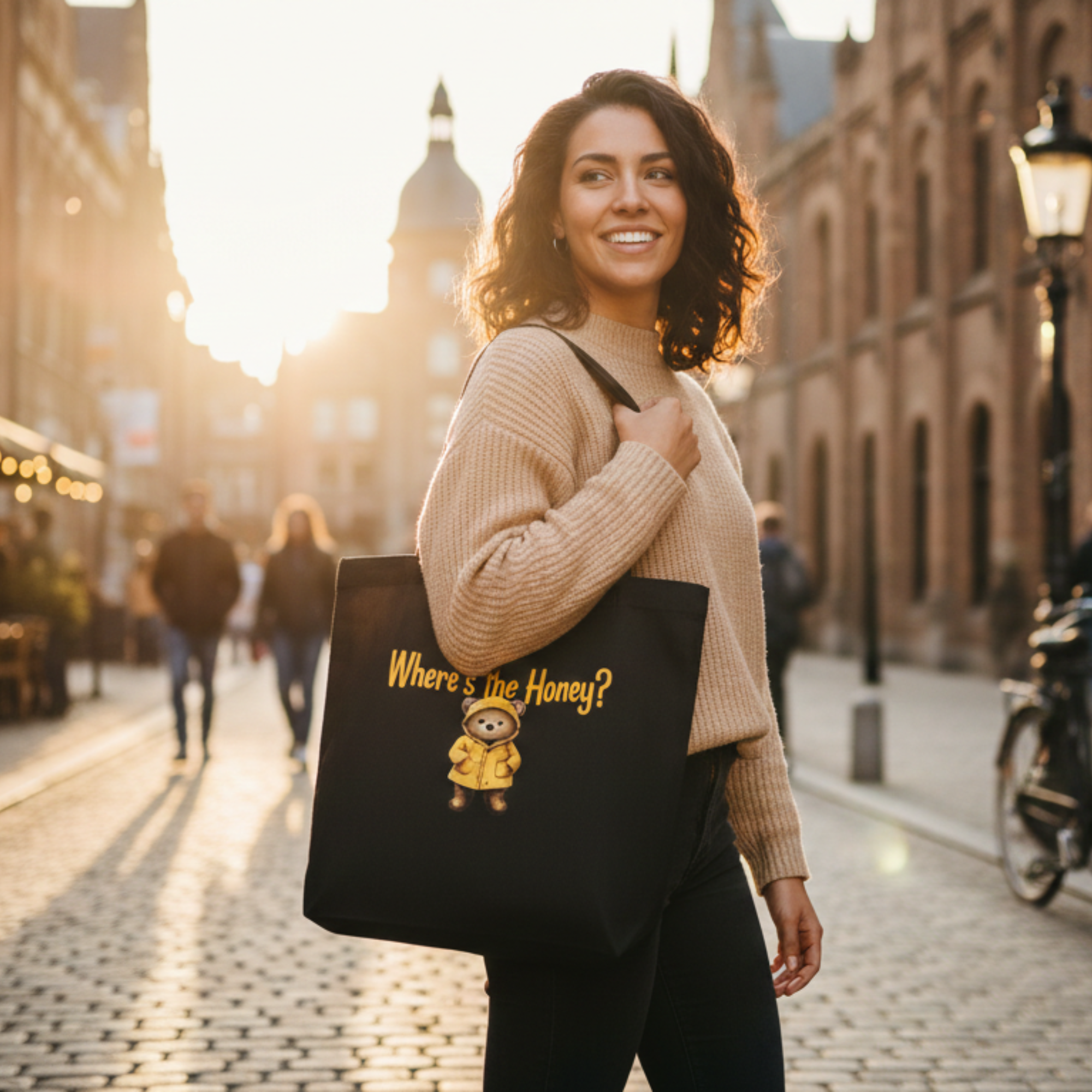 A smiling woman carrying a black eco-friendly tote bag featuring a whimsical bear in a yellow raincoat with the text 'Where's the Honey?', walking down a sunlit European-style cobblestone street.