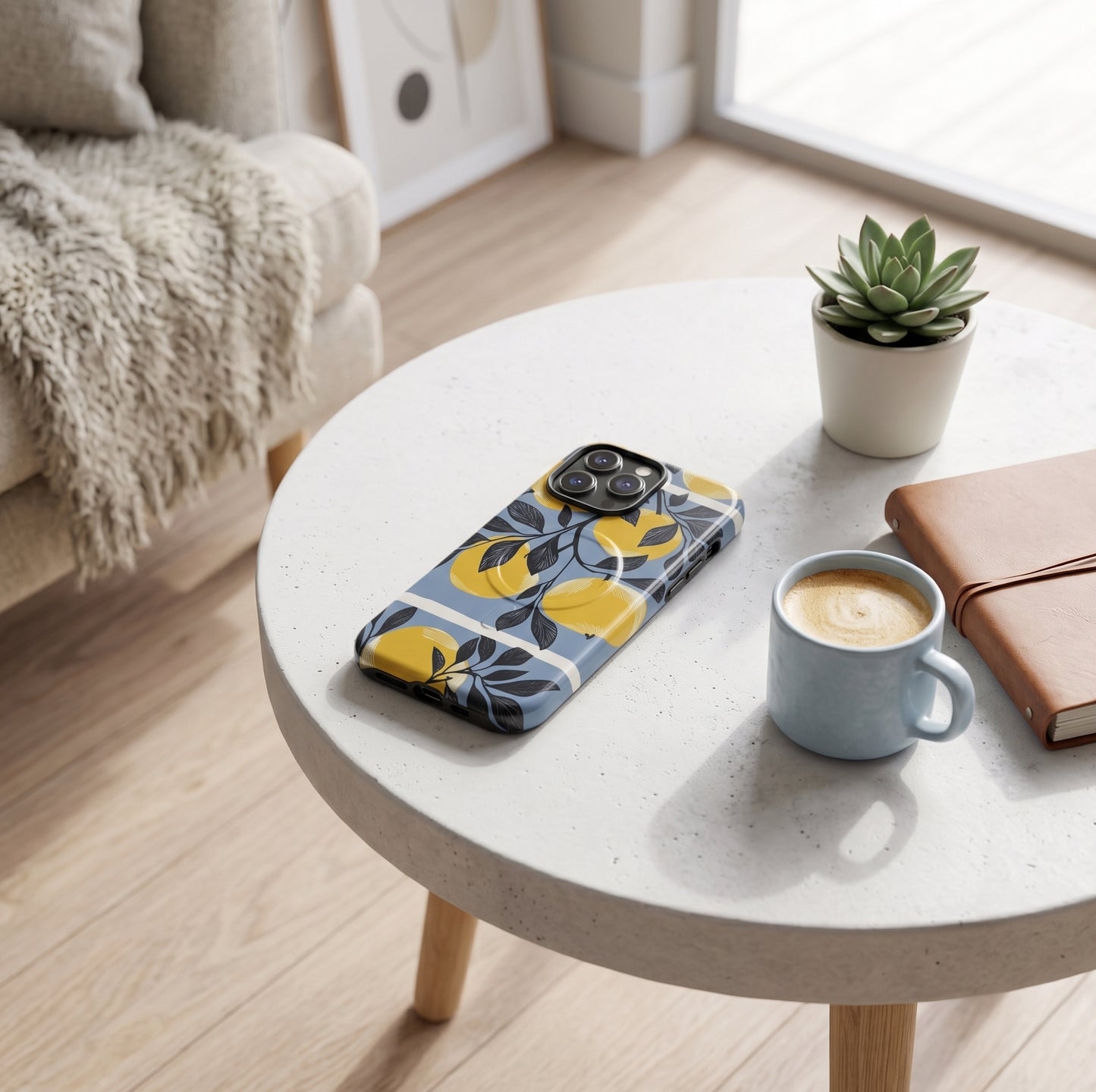 A MagSafe iPhone case with yellow apples and blue floral pattern is displayed on a round white coffee table in a cozy living room setting, alongside a cup of coffee, a notebook, and a small potted succulent.