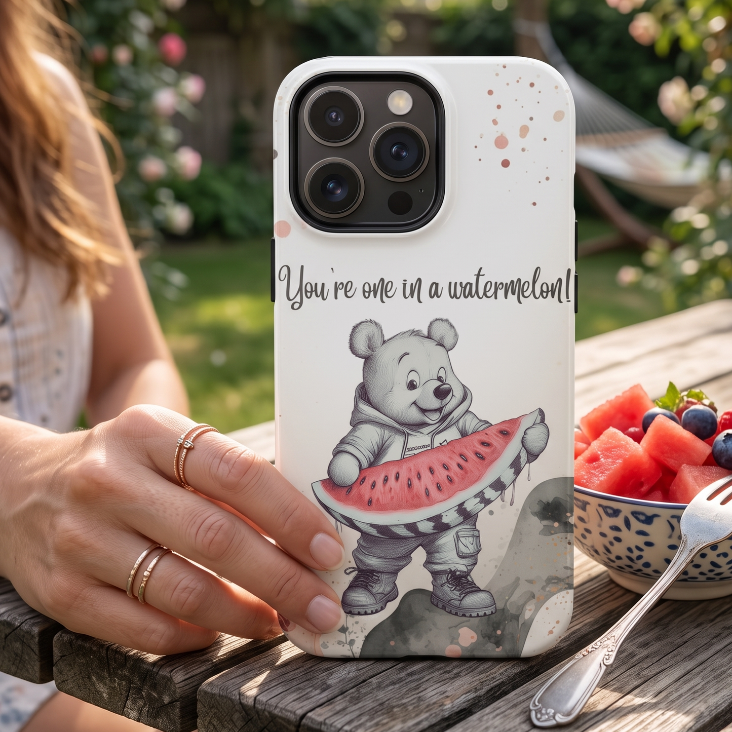 A white phone case featuring Winnie the Pooh holding a watermelon slice with the text 'You're one in a watermelon!' is held by a person at an outdoor wooden table with a bowl of fruit in the background.