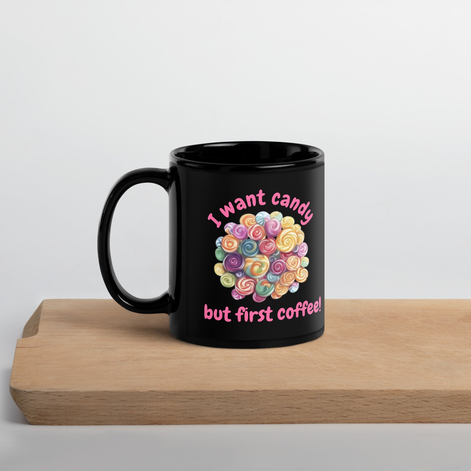 A black ceramic mug with a colorful heart-shaped lollipop graphic and the text 'I want candy but first coffee!' sitting on a light wooden board against a white background.