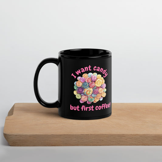 A black ceramic mug with a colorful heart-shaped lollipop graphic and the text 'I want candy but first coffee!' sitting on a light wooden board against a white background.