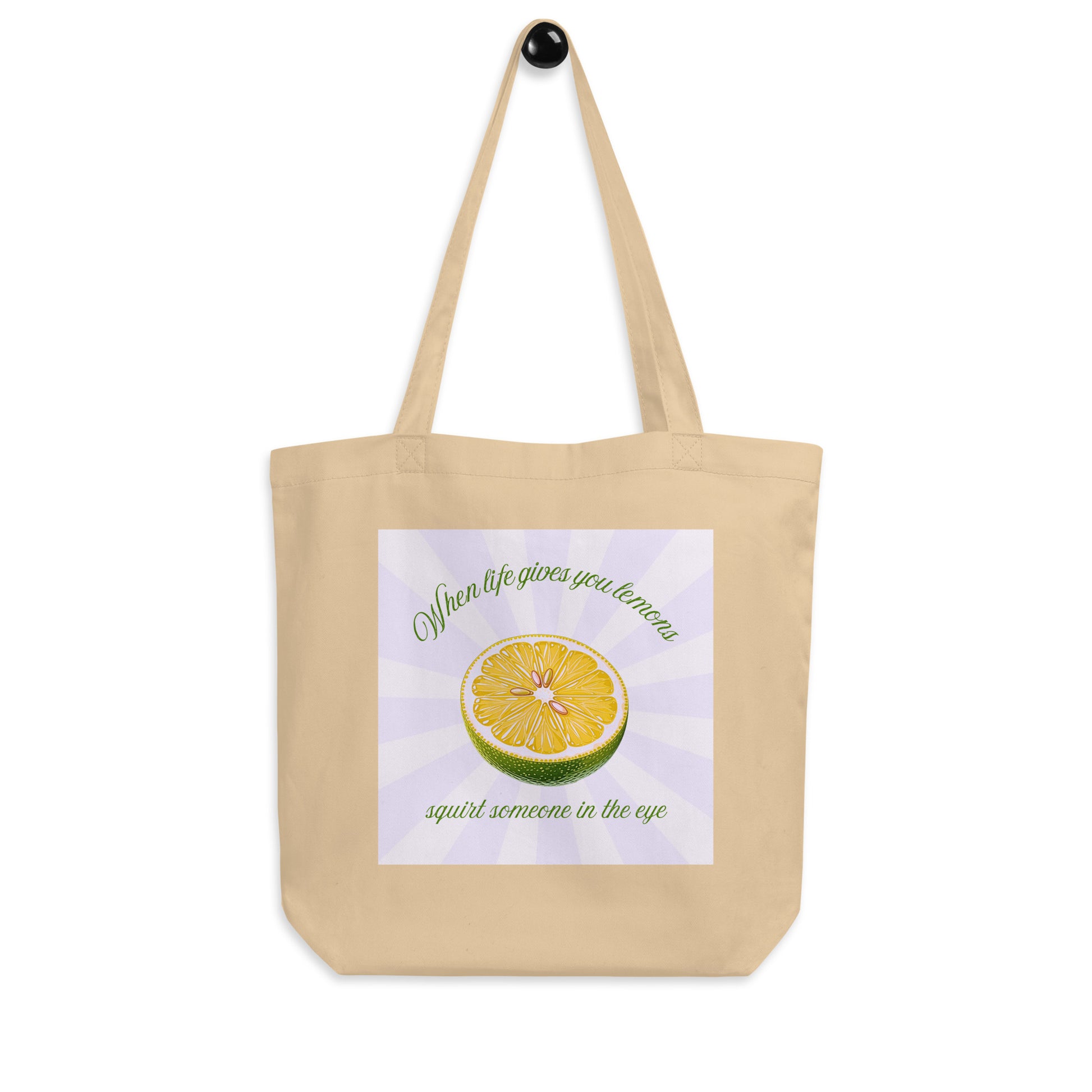 A beige canvas tote bag with a black hanging loop, featuring a printed graphic of a sliced lime with the text 'When life gives you lemons squirt someone in the eye' by Sweet & Savage on a white background.