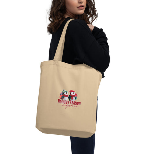 Beige eco tote bag with 'Holiday Season' design featuring two penguins holding hands being held by a person on a white background. 