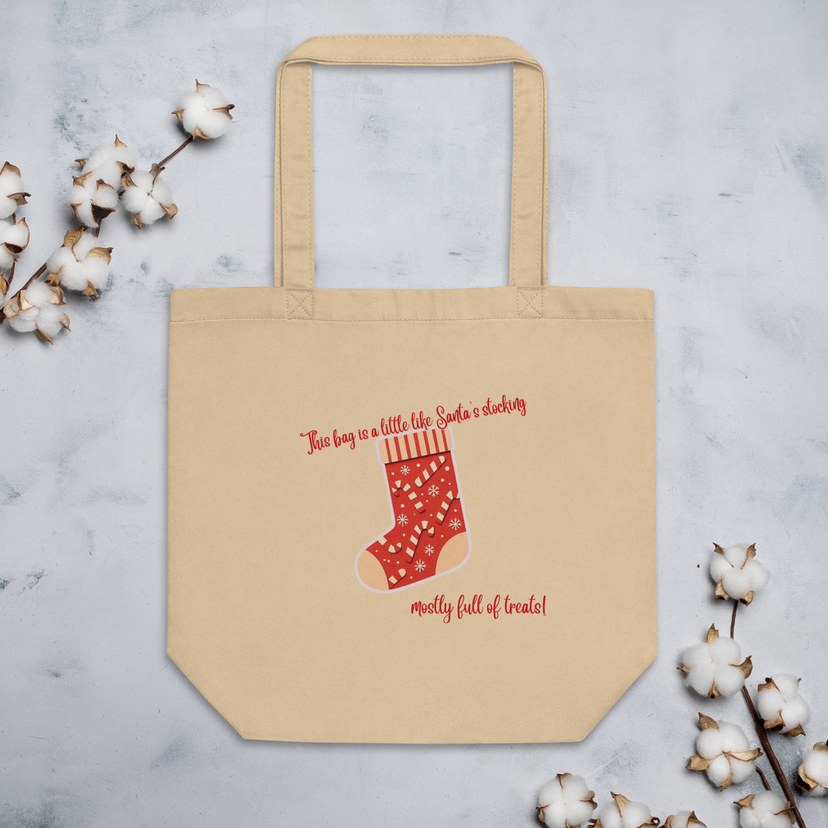A beige canvas tote bag with a red Christmas stocking graphic and the text 'This bag is a little like Santa's stocking, mostly full of treats!' on a light gray marble background with cotton stems in the corners.