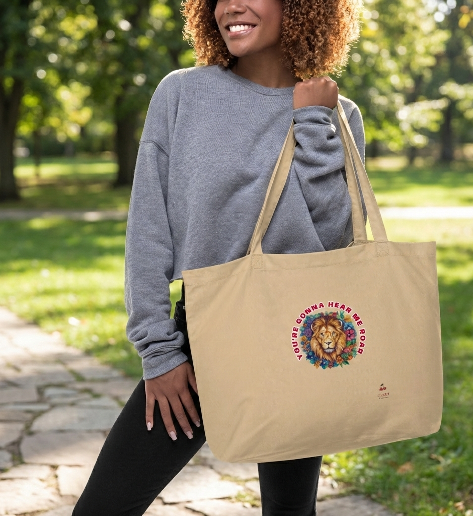 person with curly hair, wearing a grey sweatshirt and black pants, smiles while holding a light brown tote bag over their shoulder. The bag features a circular graphic with a lion's head and a colorful mane, surrounded by the red text "YOU'RE GONNA HEAR ME ROAR." They are standing on a stone path in a park with green grass and trees in the background.
