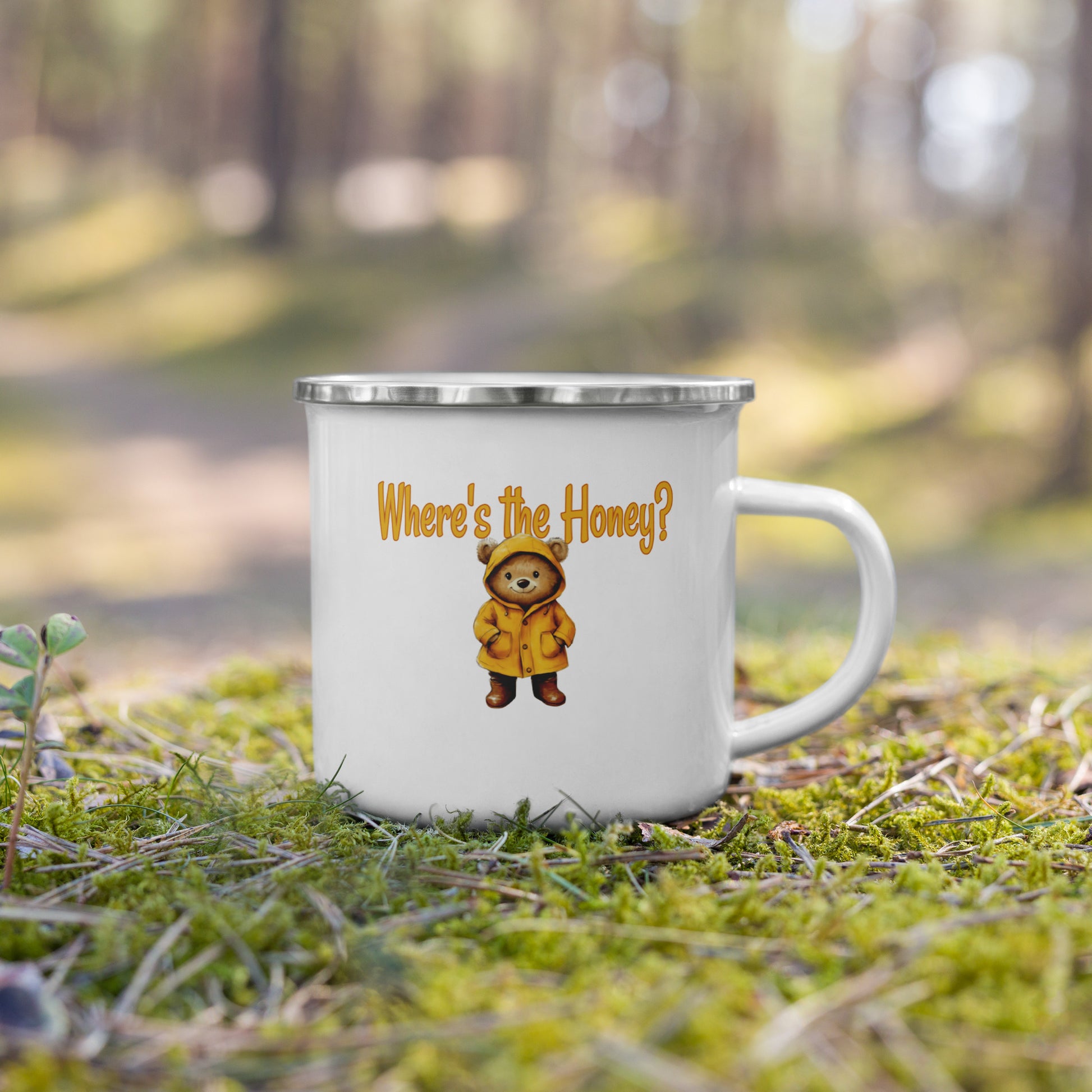 A white enamel-style mug with a silver rim sits on mossy ground outdoors, with a blurred forest background. The mug is printed with the phrase "Where's the Honey?" in orange above a small cartoon bear in a yellow raincoat.