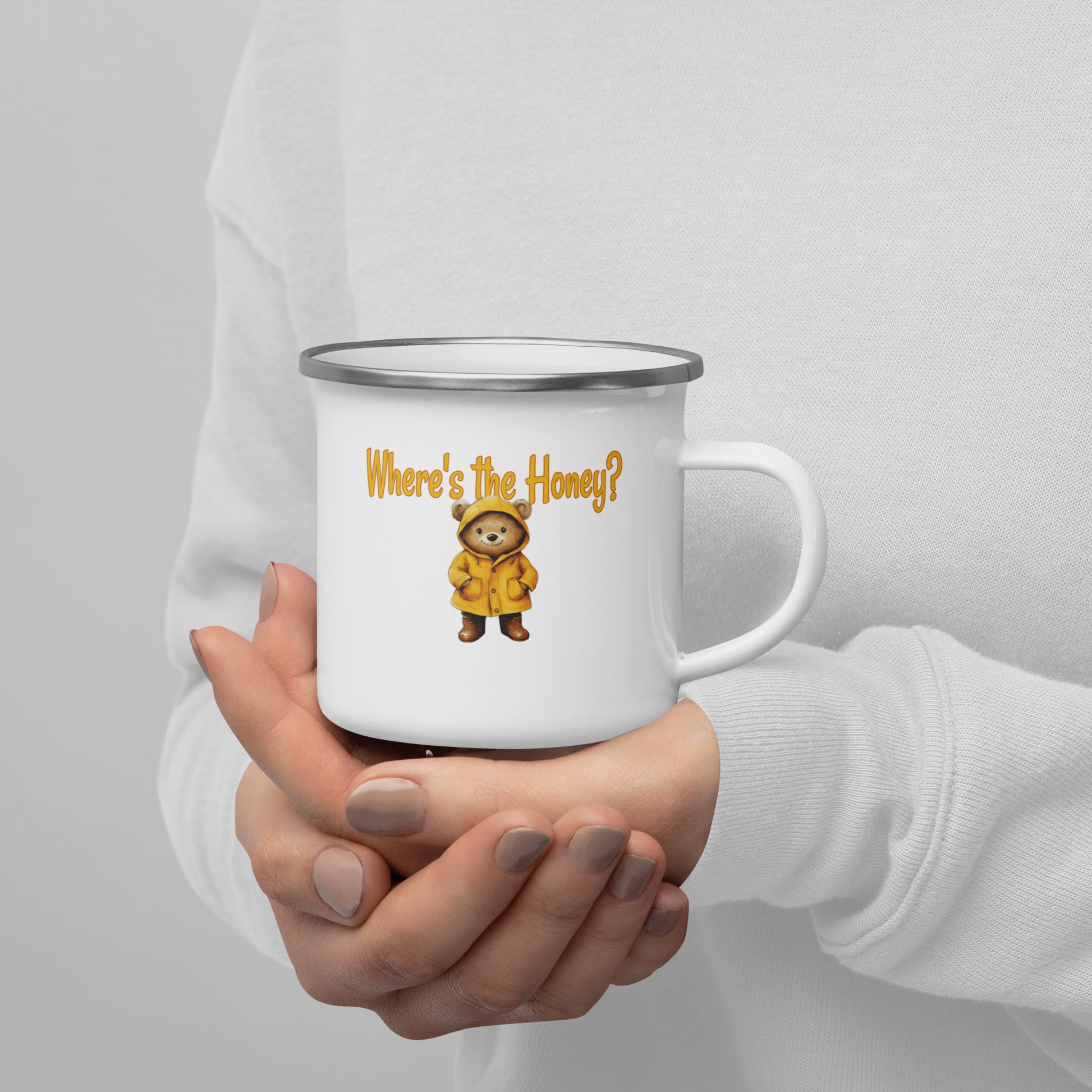 A person in a white sweatshirt holds a white enamel-style mug with a silver rim. The mug has a yellow text reading 'Where's the Honey?' and a small cartoon bear wearing a yellow raincoat.