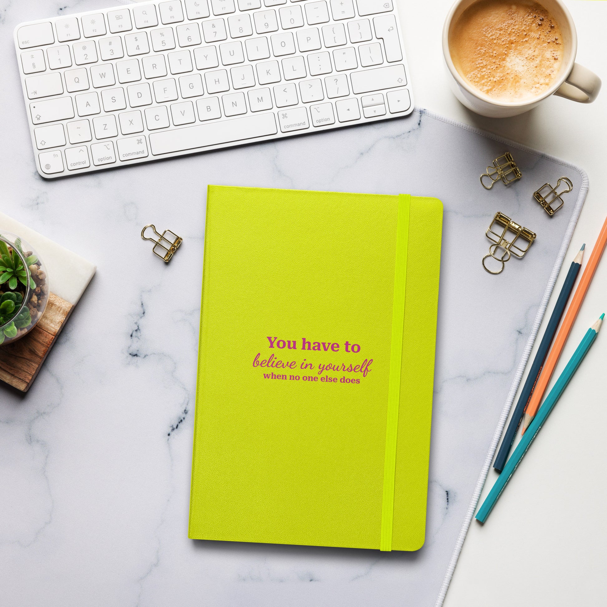 Lime green hardcover notebook with the text "You have to believe in yourself" on a marble desk with a keyboard and coffee.