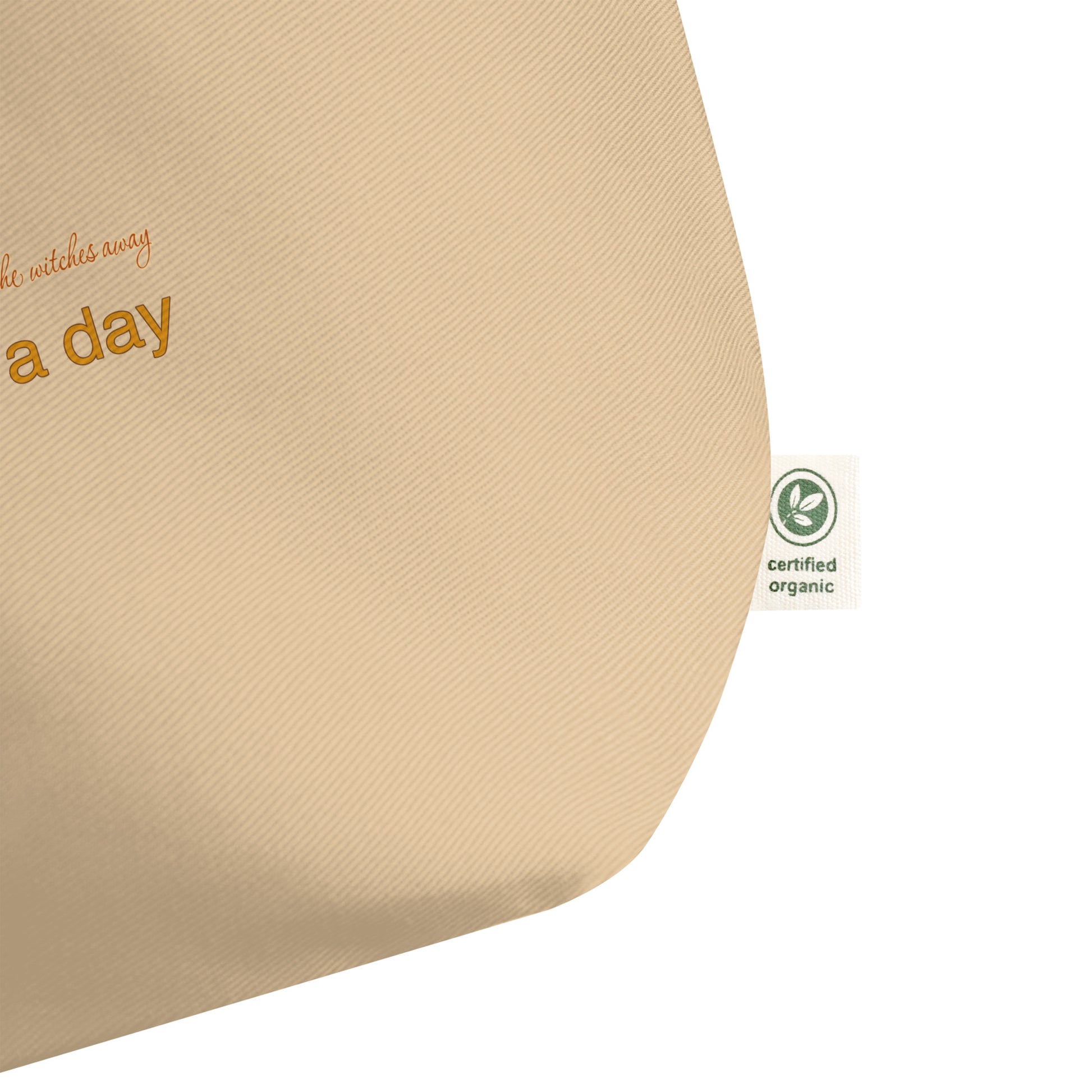 Close-up of a beige canvas tote bag corner showing partial orange text and a small green Certified Organic tag on a white background.