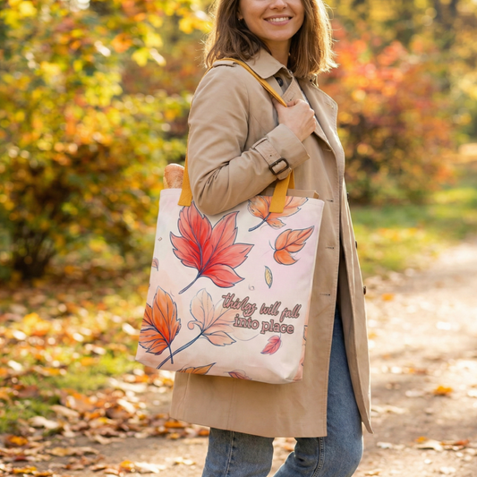 A woman dressed in a long coat holding a tote bag with autumn leaf design and text "Things will fall into place" in an outdoor setting.
