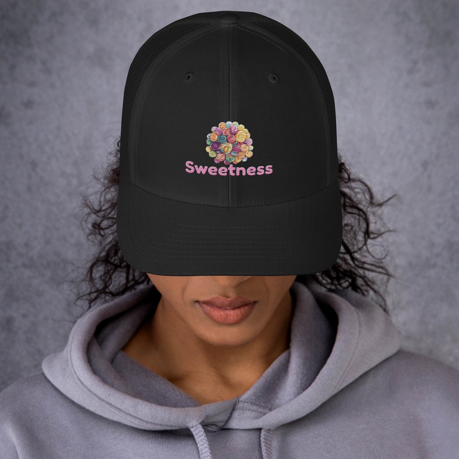 Person wearing a black cap with 'Sweetness' text and colorful candies  against a gray background