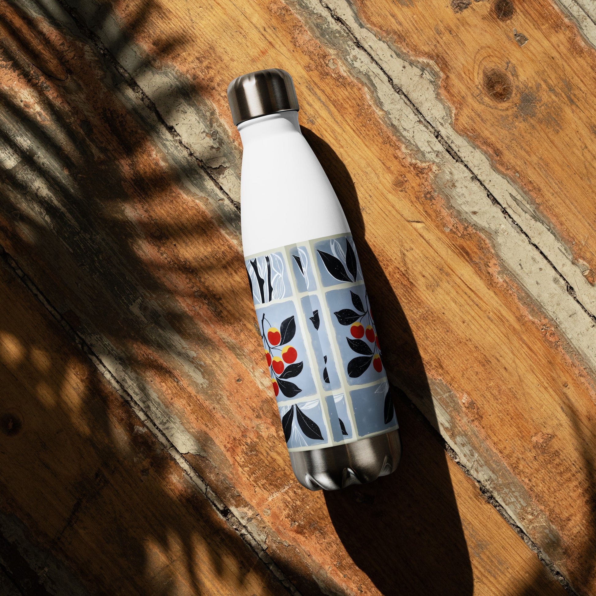 A white insulated water bottle with a blue floral pattern and red berry accents, resting diagonally on a rustic wooden surface with dappled sunlight and shadows.