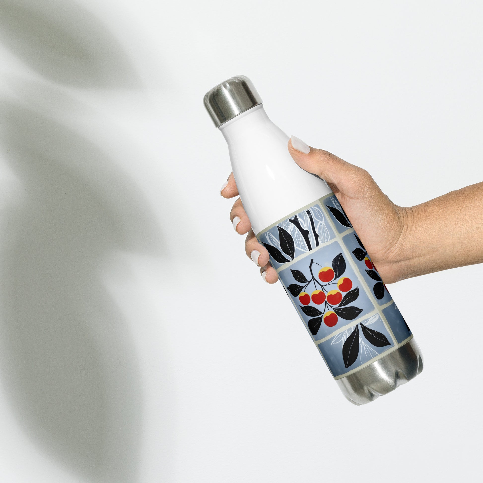 A hand holding a white insulated water bottle with a blue, black, and red cherry pattern on the lower half, against a light gray background with soft shadows.