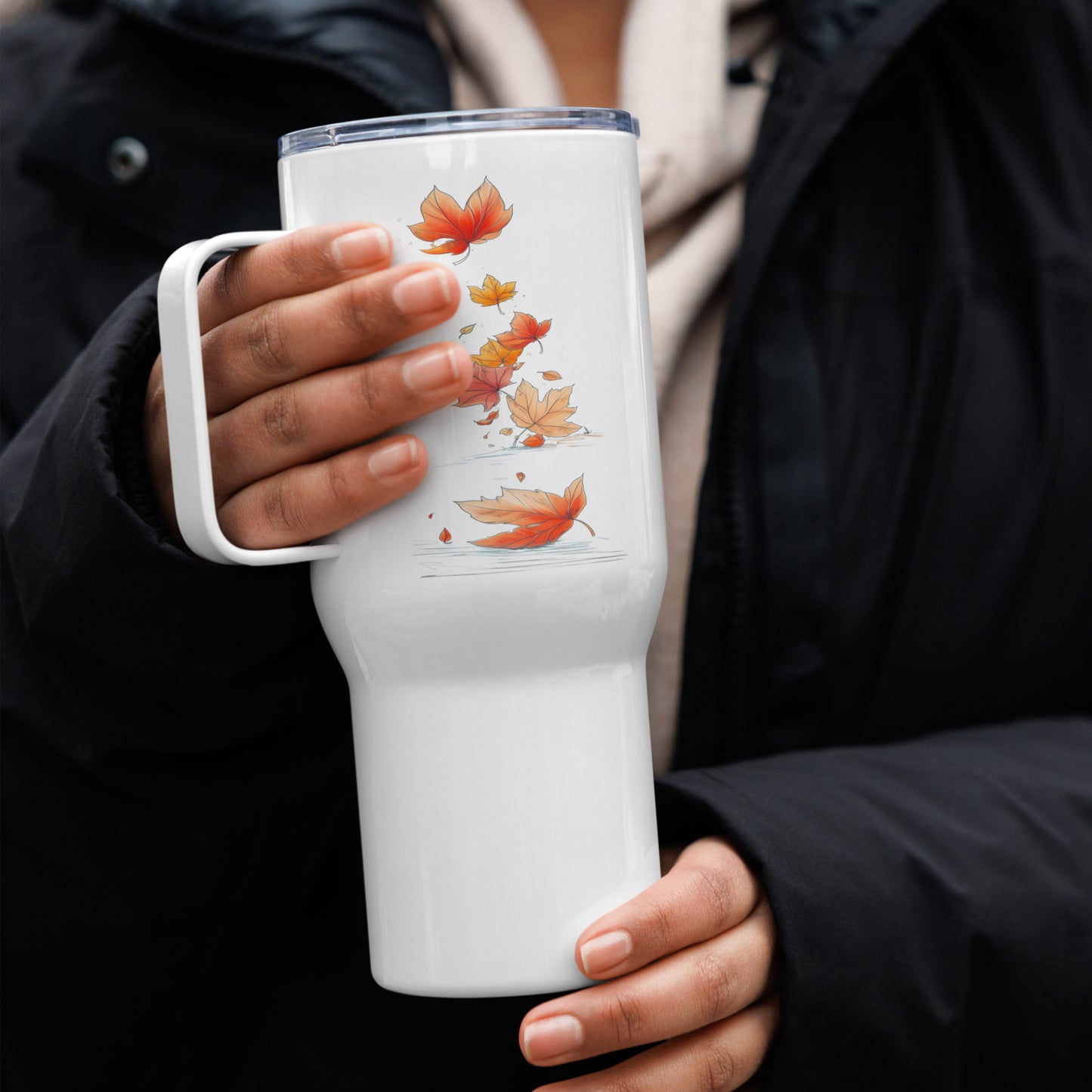 A person in a black jacket holds a white insulated tumbler with a clear lid and a printed design of orange and yellow autumn leaves.