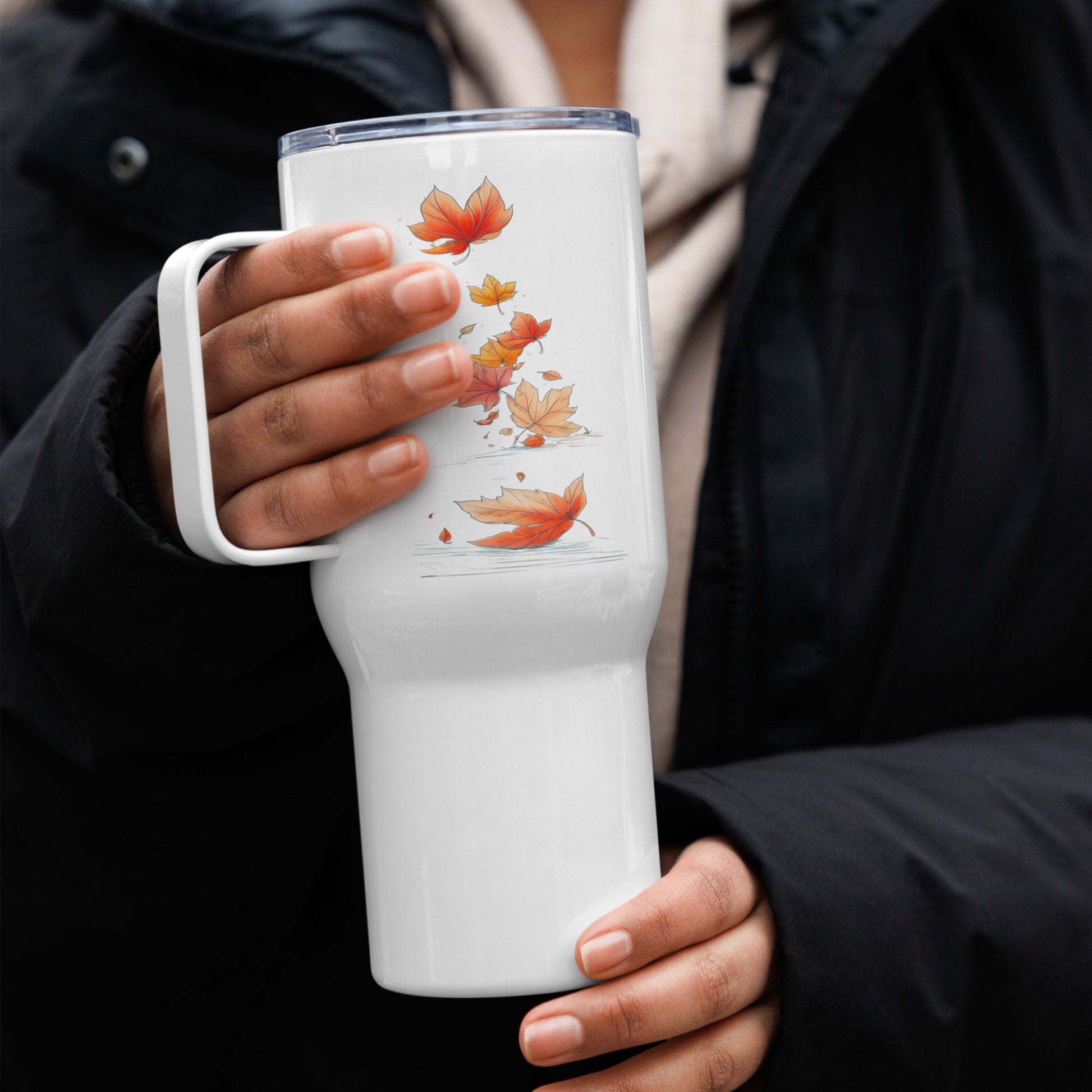 A person in a black jacket holds a white insulated tumbler with a clear lid and a printed design of orange and yellow autumn leaves.