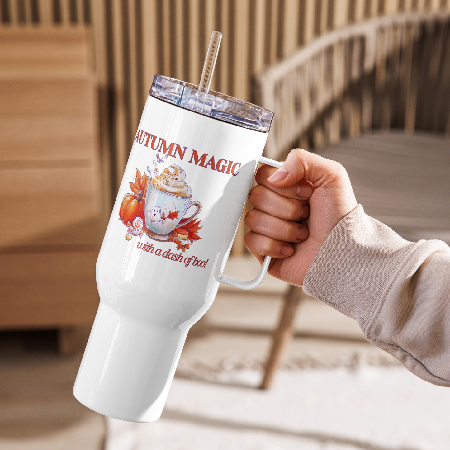 A hand holding a white insulated tumbler with a clear lid and straw. The tumbler features an autumn-themed graphic with the text 'AUTUMN MAGIC with a dash of boo!' and illustrations of a mug, hot chocolate, pumpkins, and fall leaves by Sweet & Savage. 