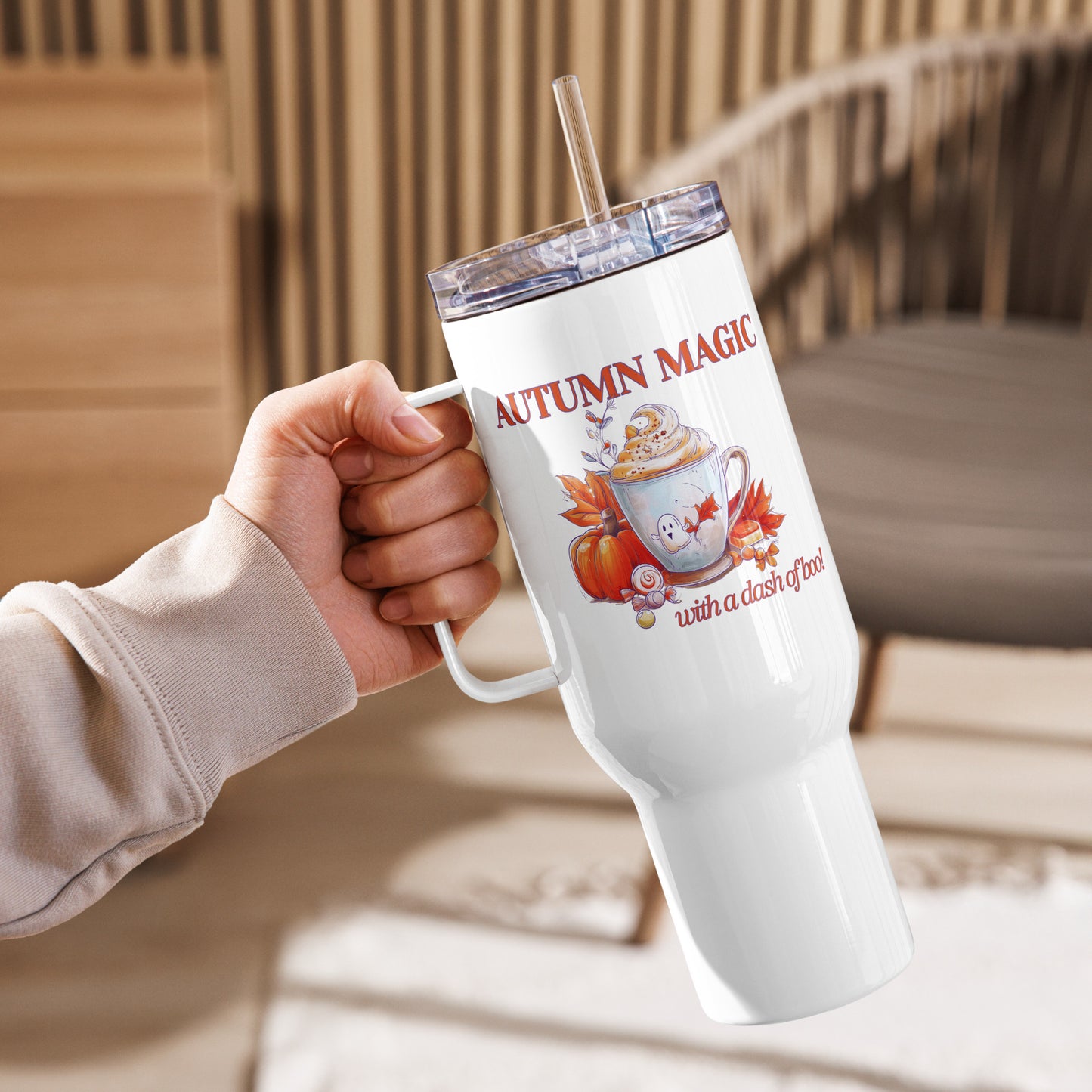 A hand holding a white insulated tumbler with a clear lid and straw. The tumbler features a printed autumn-themed graphic with the words 'AUTUMN MAGIC' and 'with a dash of boo!' by Sweet & Savage. 