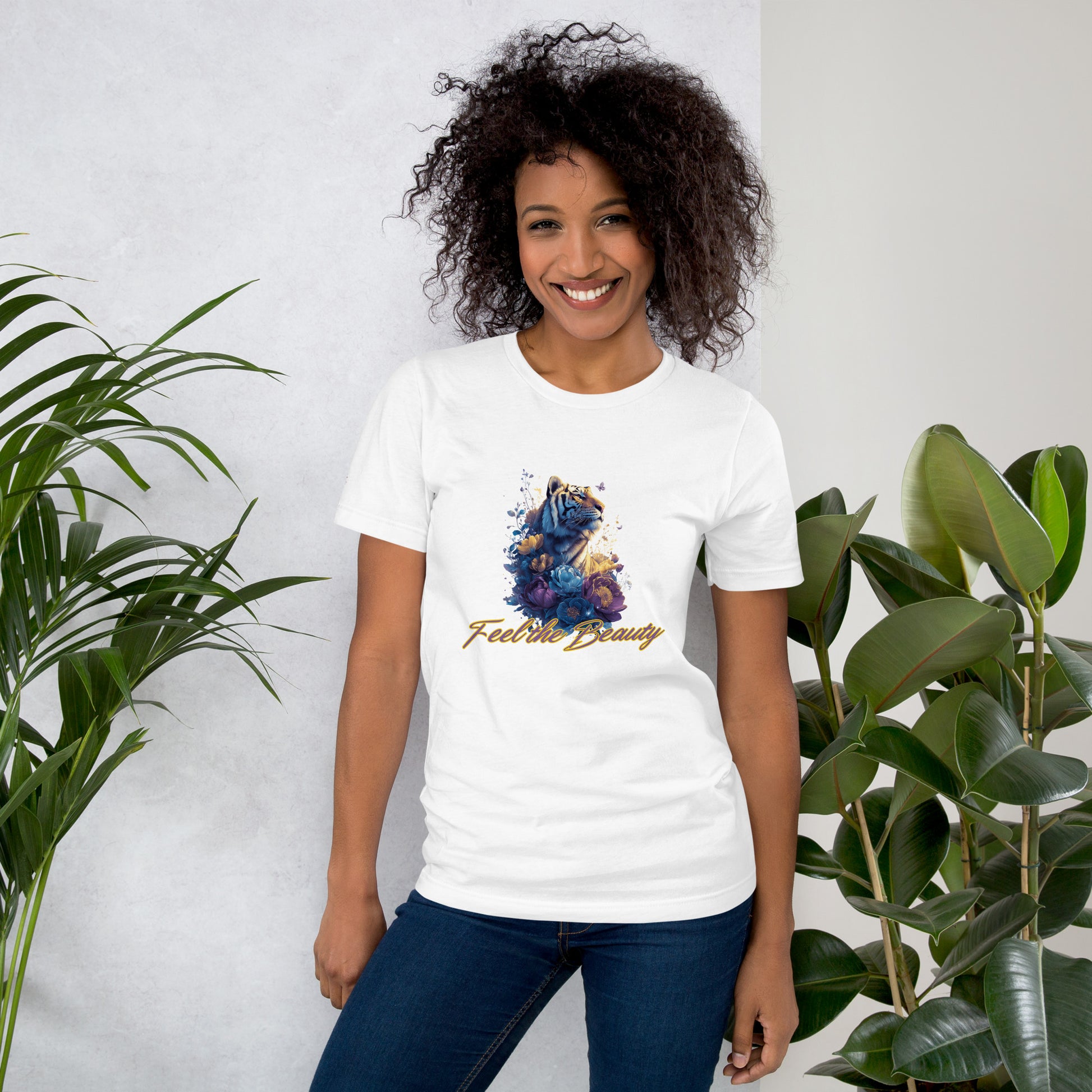 Woman wearing a white t-shirt with a colorful design of a tiger and a text that says 'Feel the Beauty'.The woman is standing between two plants.