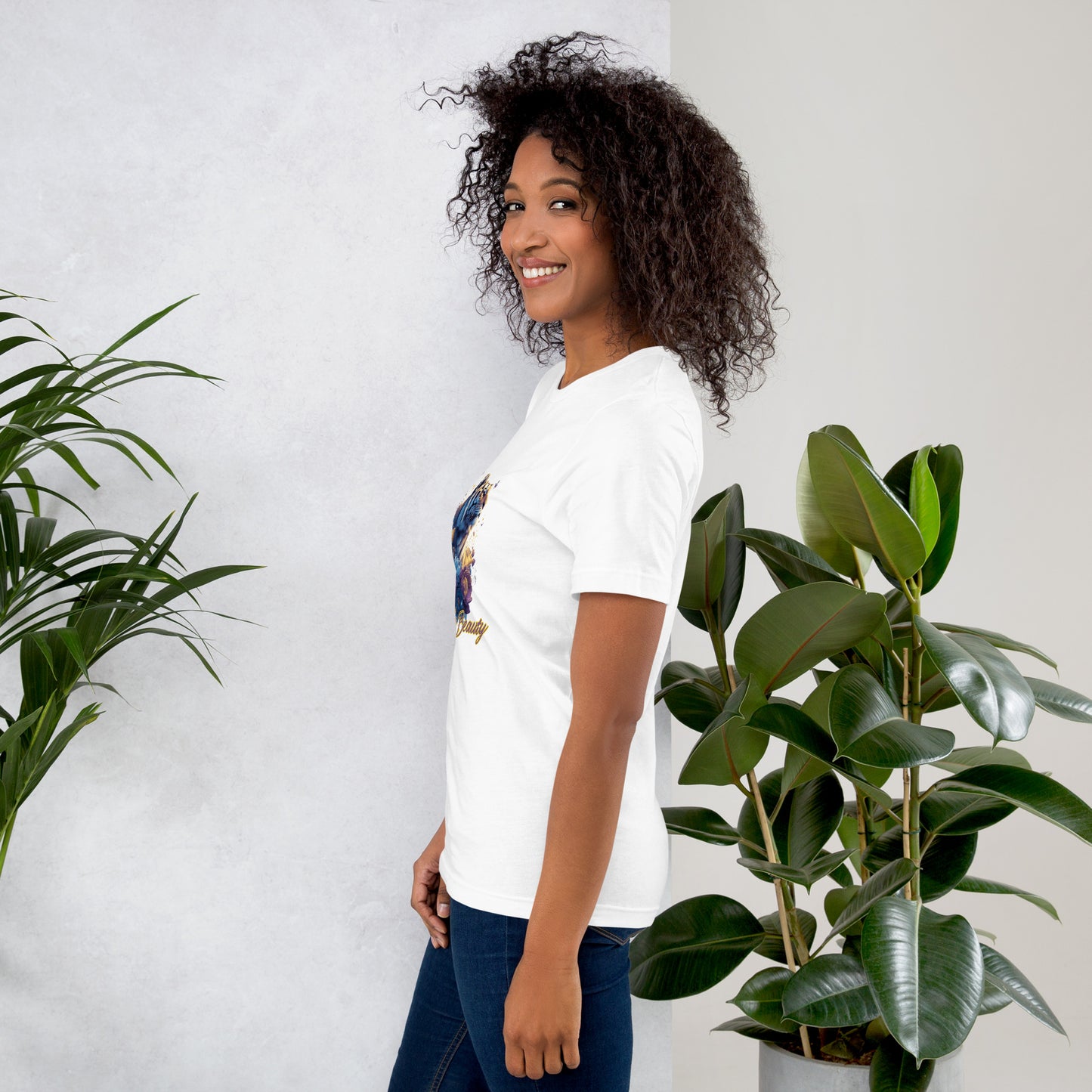 Woman wearing a white t-shirt with a colorful graphic design, standing between two potted plants against a light gray wall.