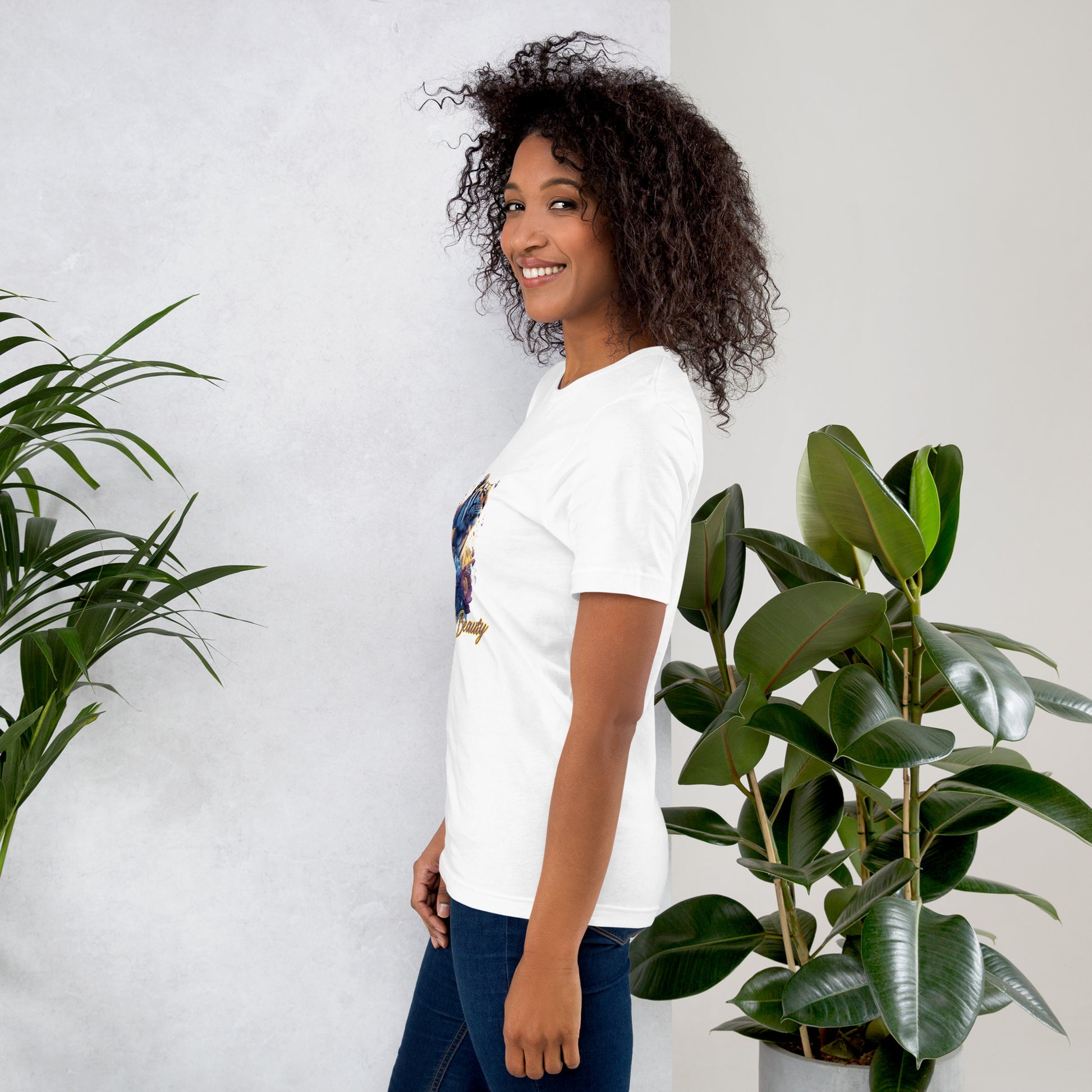 Woman wearing a white t-shirt with a colorful graphic design, standing between two potted plants against a light gray wall.